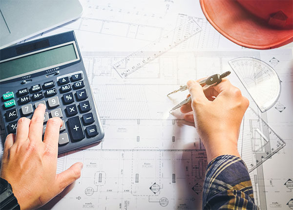 Fotografía de las manos de una persona con una calculadora, reglas y compás sobre planos
