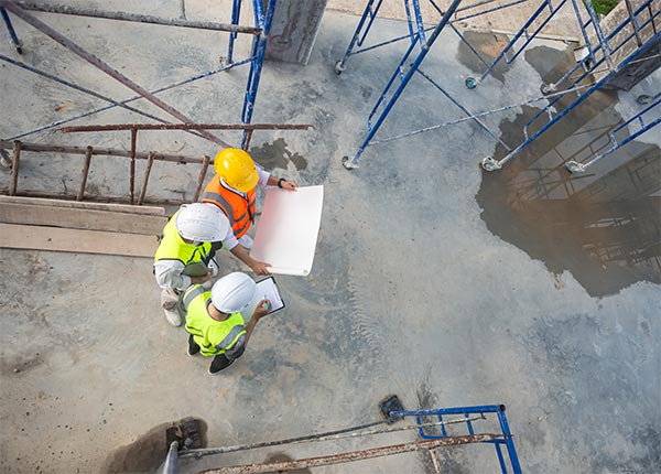 Fotografía de tres hombres con chaleco y casco en una obra consultando un plano con cemento en el suelo y andamios