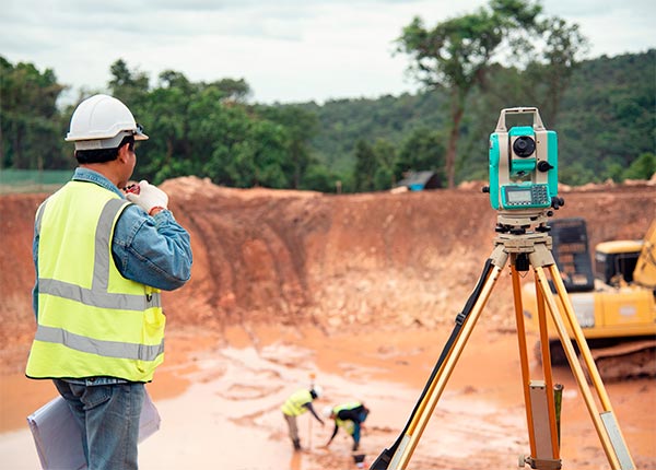 Fotografía de un hombre en una obra con chaleco y casco supervisando el suelo con dos hombres al fondo trabajando
