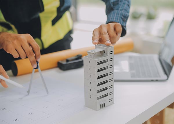 Fotografía de una persona con un chaleco de obra trabajando sobre una mesa con planos, una maqueta de edificio y un compás